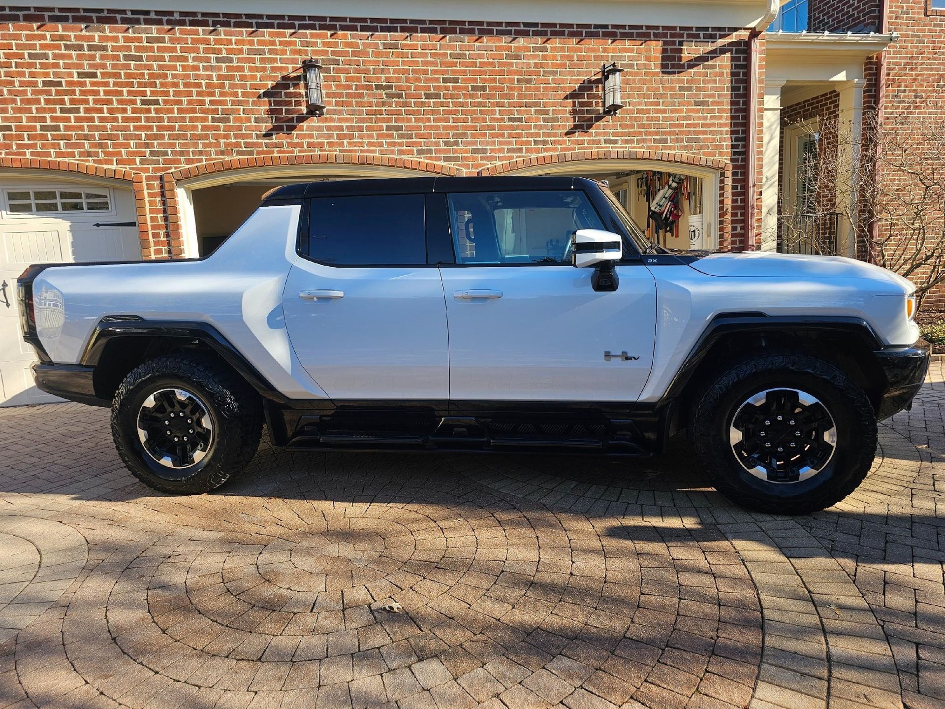 White GMC Hummer EV pickup truck parked in front of a brick house with a paved driveway.