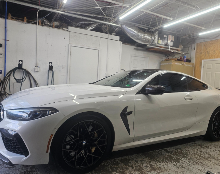 White BMW coupe with black wheels parked inside a garage, next to a wall with equipment.