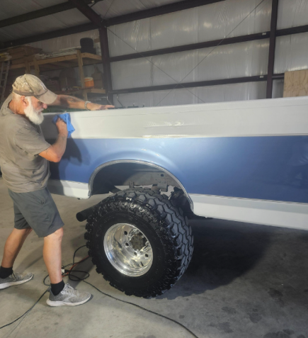Man wiping the side of a blue and white pickup truck with oversized tires inside a garage.