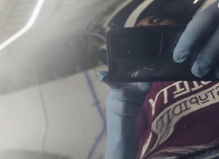 Reflection in a shiny surface, showing a person wearing blue gloves holding a phone. A red and white shirt is visible.