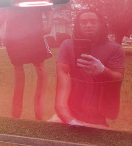 Man in a gray shirt takes a selfie outdoors in front of a red surface. Another person is reflected.