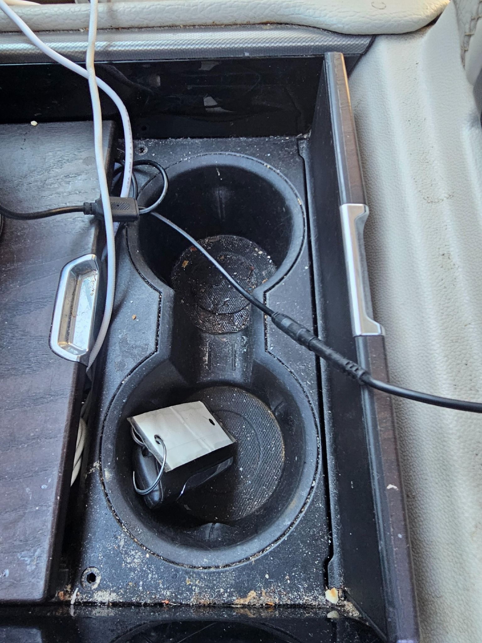 Center console of a car with two cup holders, debris, and a device connected to cables.
