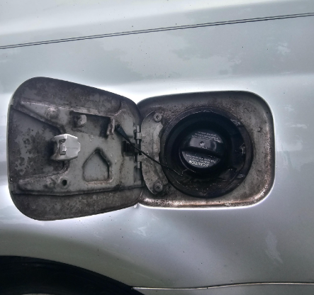 Open fuel door of a silver car, revealing a black fuel cap and dirty compartment.