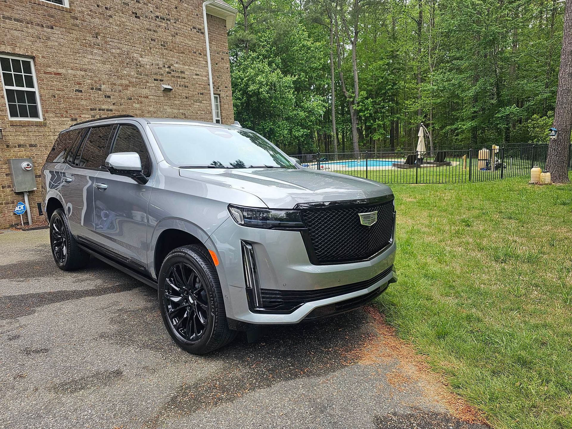 Silver Cadillac Escalade SUV parked on a gravel driveway in front of a brick building and grassy yard.