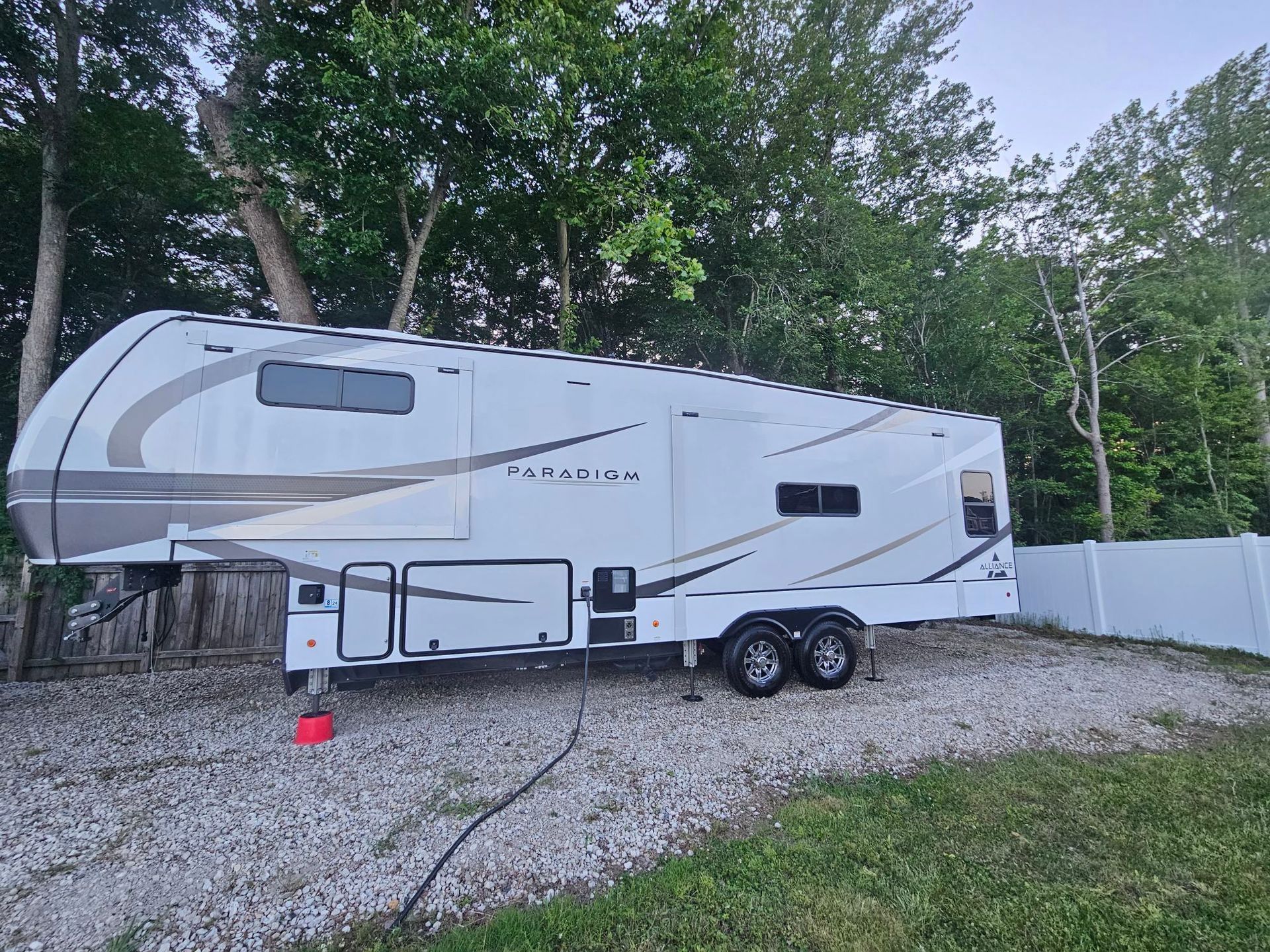 White RV parked on gravel next to a white fence and trees, power cord plugged in.