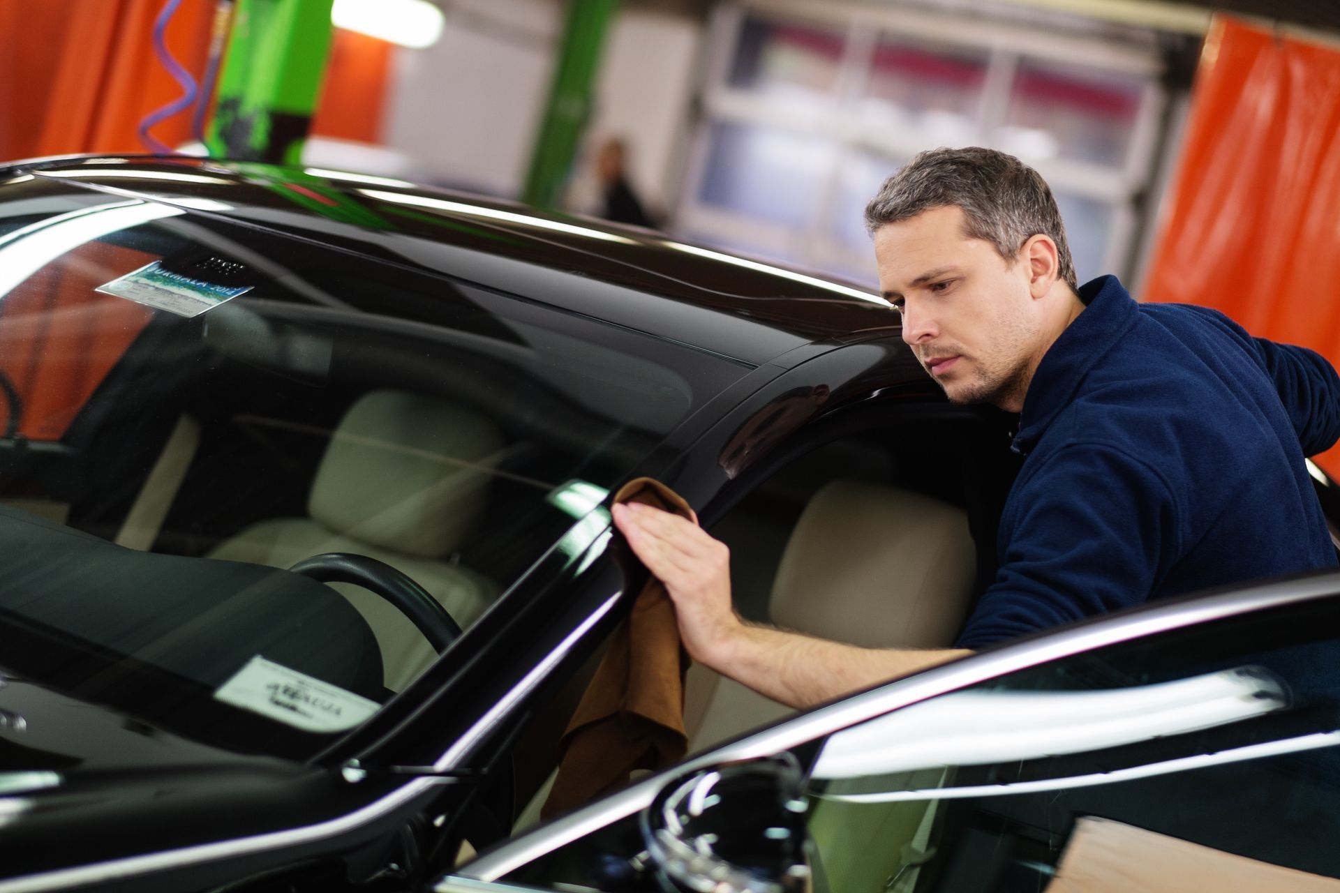 Man wiping a black car's windshield with a cloth inside a garage.