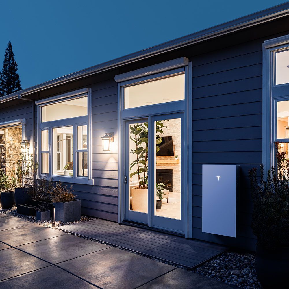 Exterior of a house at night, featuring a Tesla Powerwall installed on the wall and lit interior.