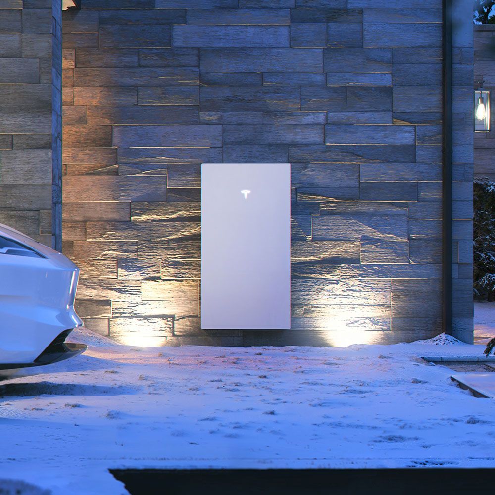 Tesla Powerwall mounted on a stone wall, lit by spotlights. A Tesla car is parked nearby in the snow.