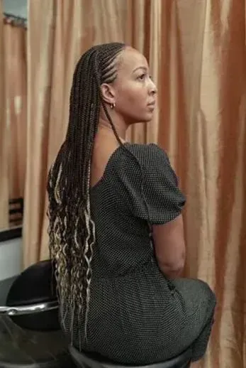 Woman with long braided hair, wearing a dark dress, sitting in front of a curtain.