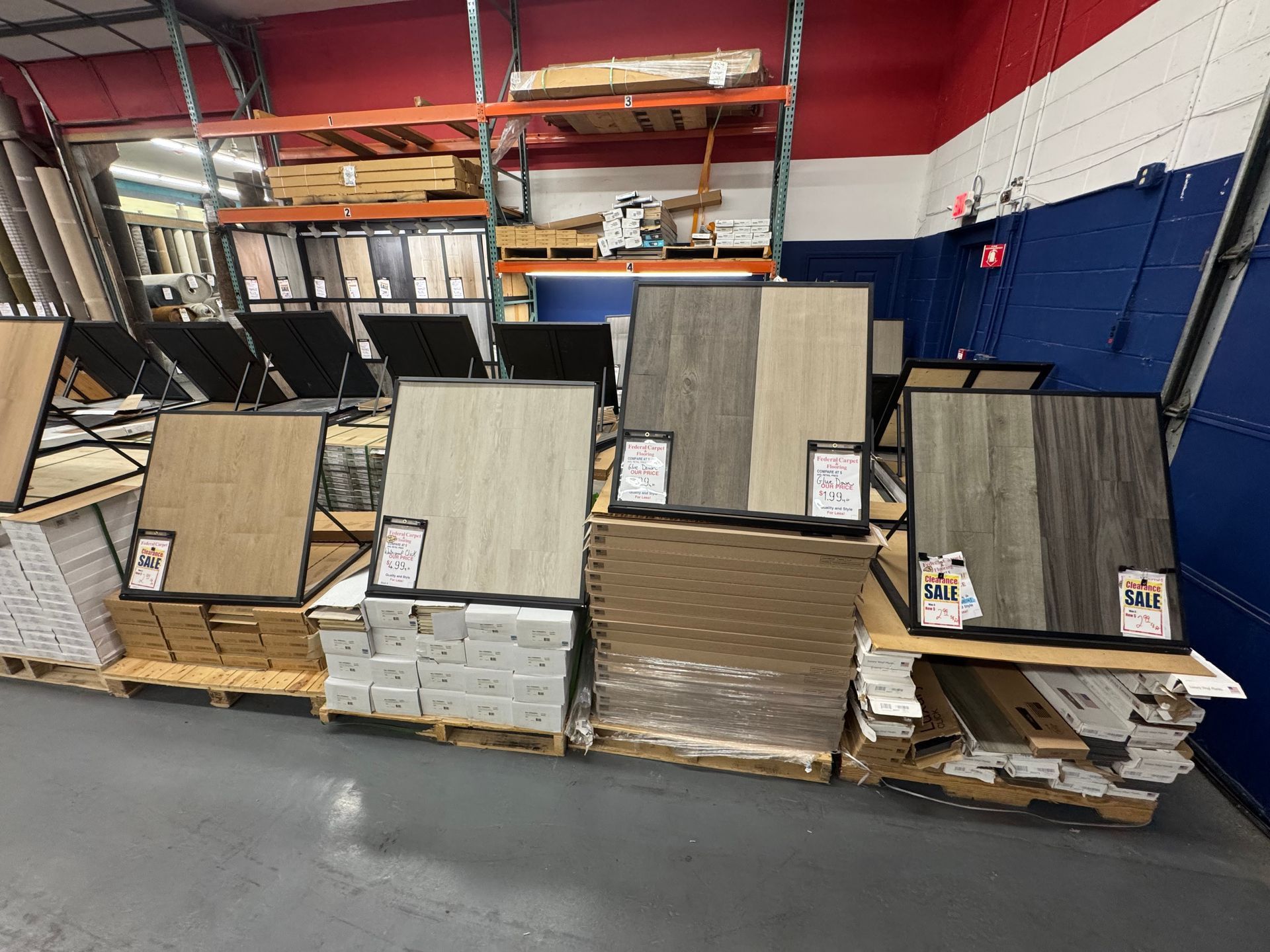 Flooring samples displayed in a warehouse-like store with wood and tile options on pallets and racks.