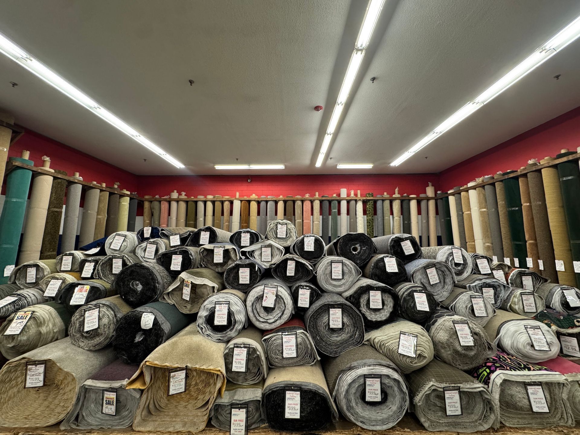 Pile of carpet rolls in a store, various colors, stacked high. Red wall background, fluorescent lighting.