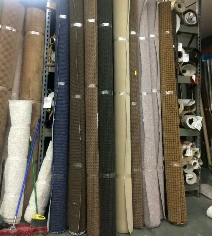 Shelves of rolled carpets in various colors, indoors. A broom leans nearby.