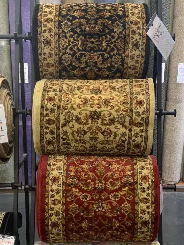 Three rolled rugs of different colors displayed on a rack. Black, beige, and red with ornate floral designs.