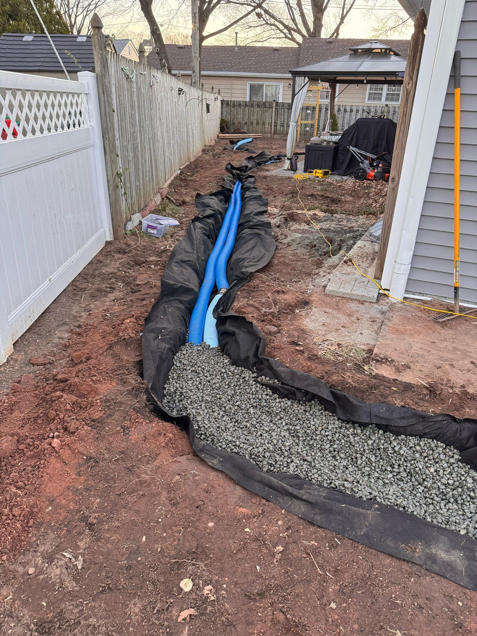 Trench with blue pipes, gravel, and black fabric alongside a house and fence.