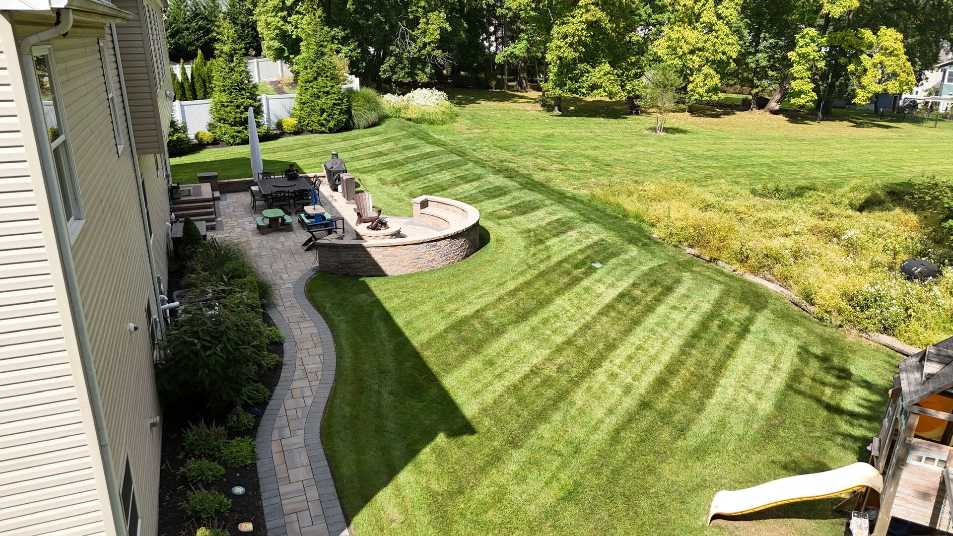 Lawn with striped pattern, patio, house exterior. Man mowing. Bright green grass, sunny day.