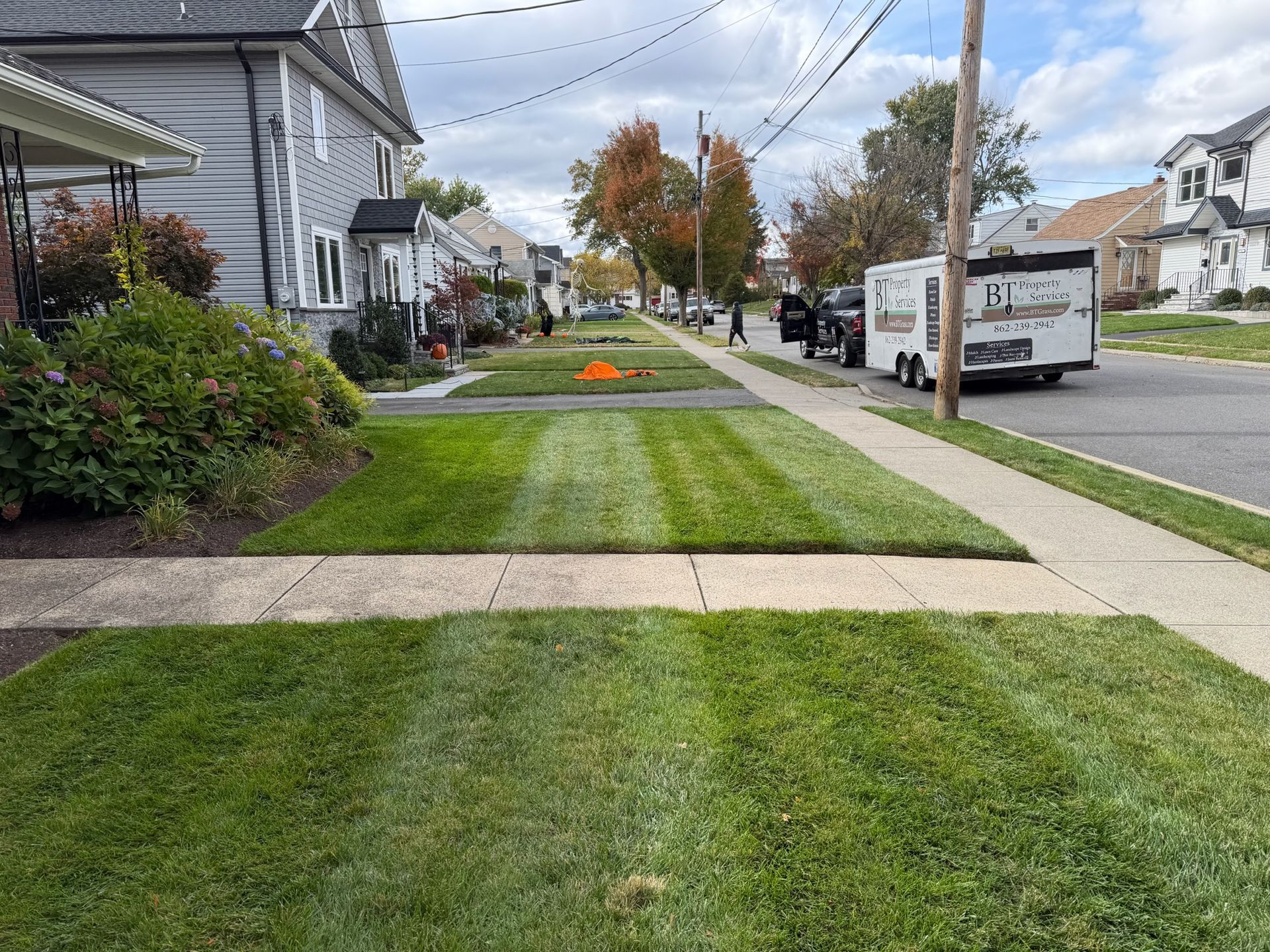 Lawncare service in progress, freshly mowed grass strips on a residential street. White trailer parked on the right.