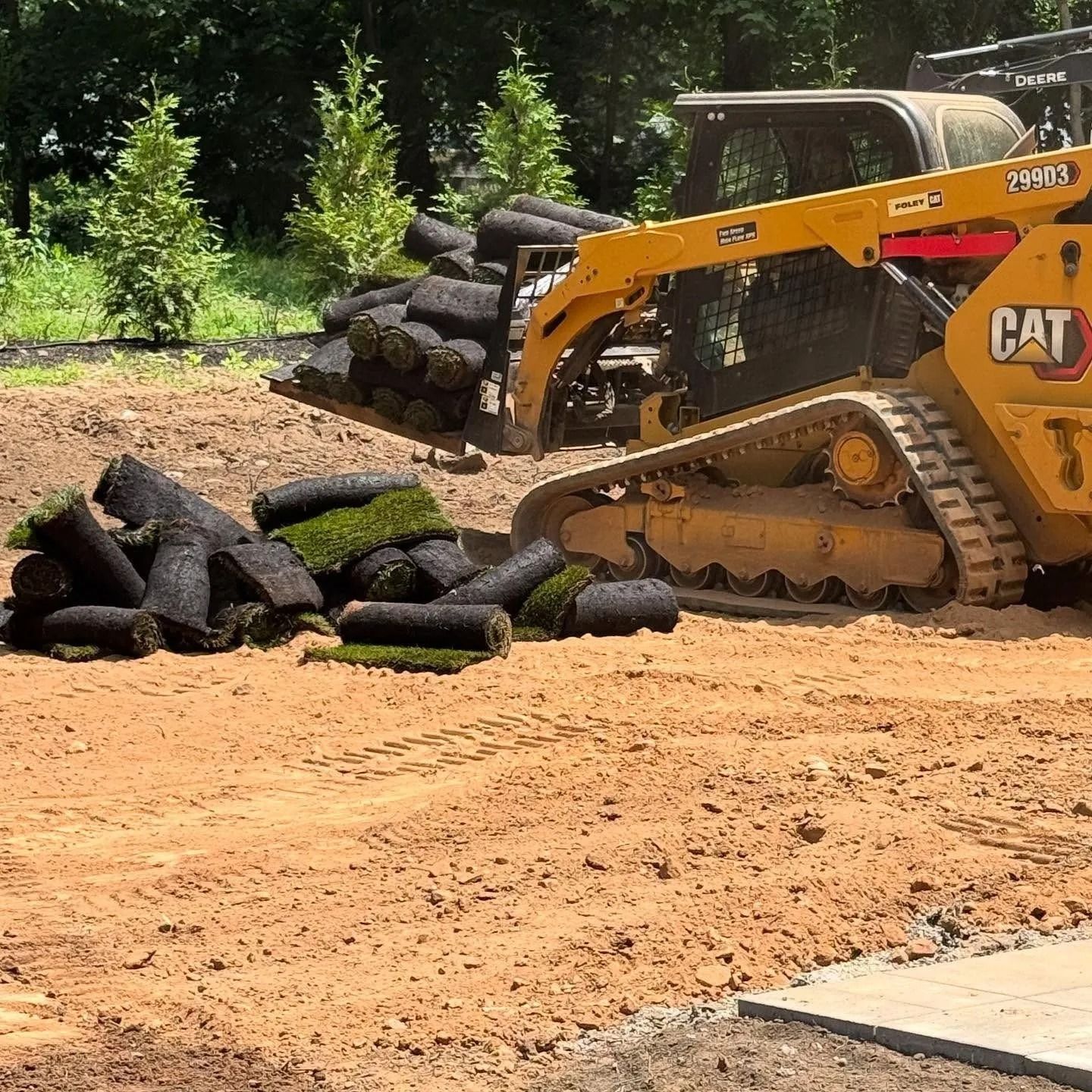 A yellow CAT skid steer unloads rolls of sod onto a dirt landscape near trees.