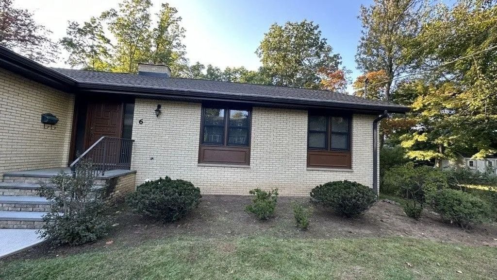 Tan brick house with dark trim and windows, a front door, and a small front yard with bushes.