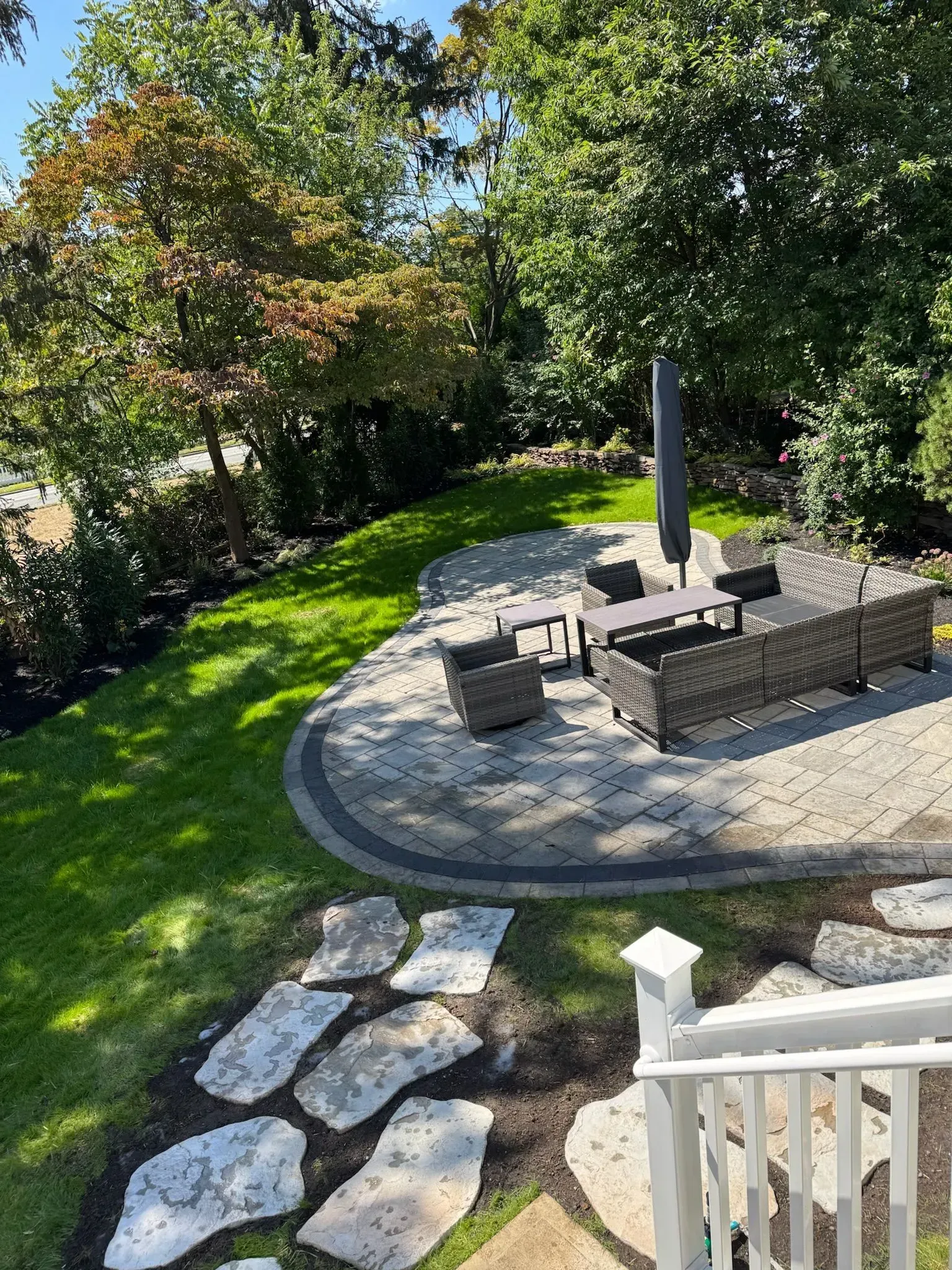 Outdoor patio with gray paving stones, furniture, and umbrella, surrounded by grass and trees.