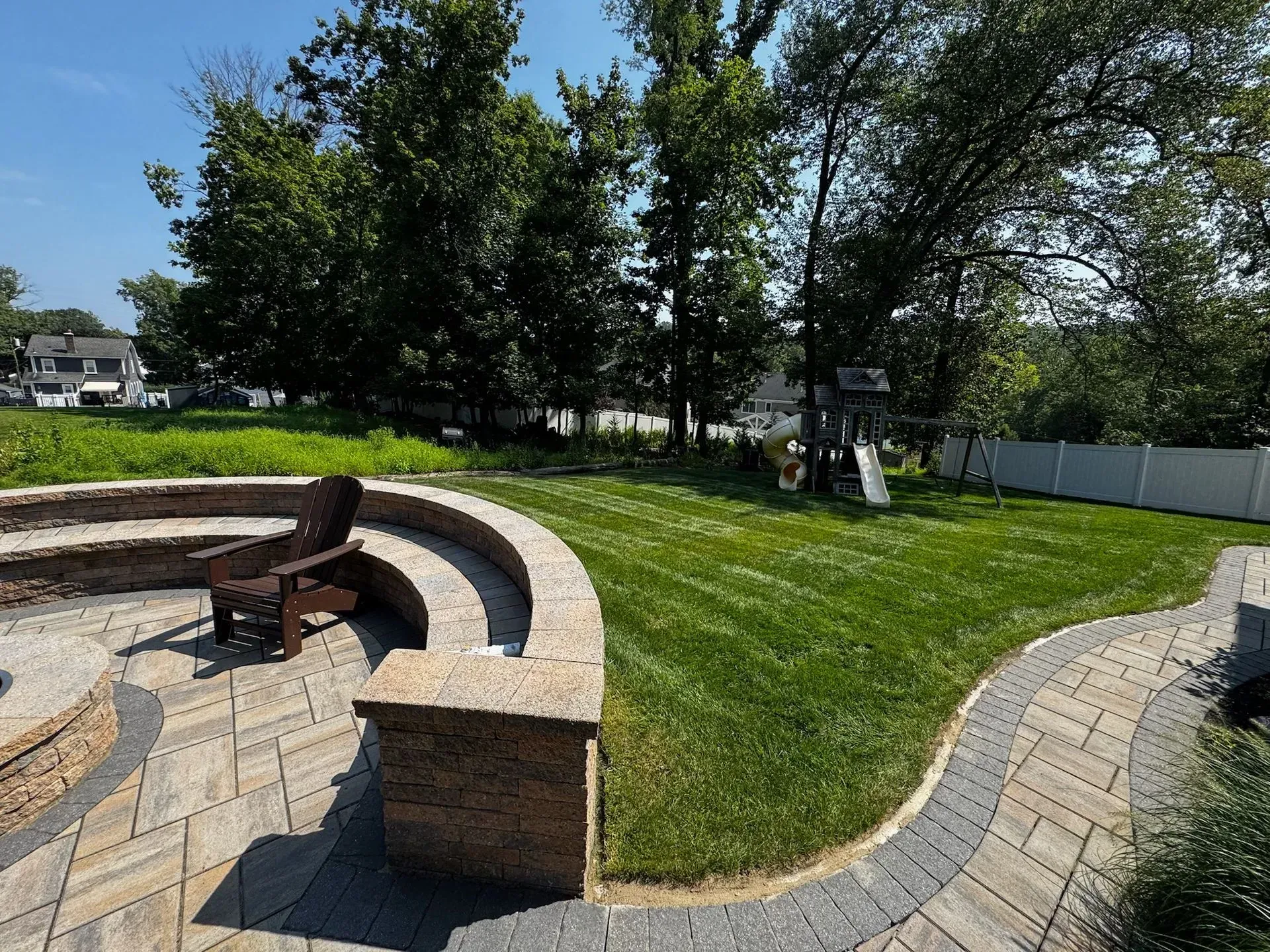 A backyard with a stone patio and seating, grass, trees, and a playset on a sunny day.