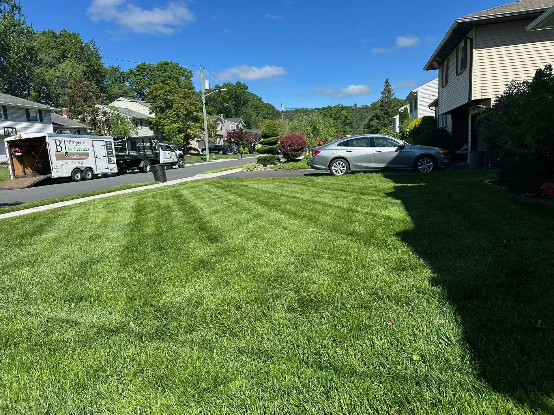 Lawn mowed with fresh stripes, a silver car parked in driveway, houses and trees in background.