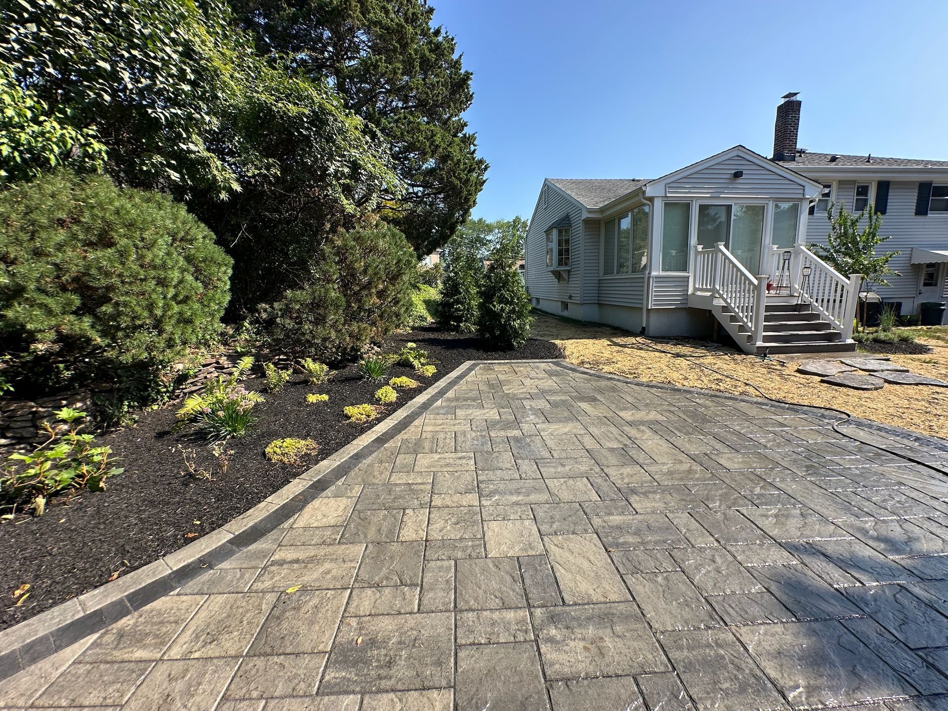 Paver driveway leading to a house with a porch. Landscaping with dark mulch and greenery. Sunny day.
