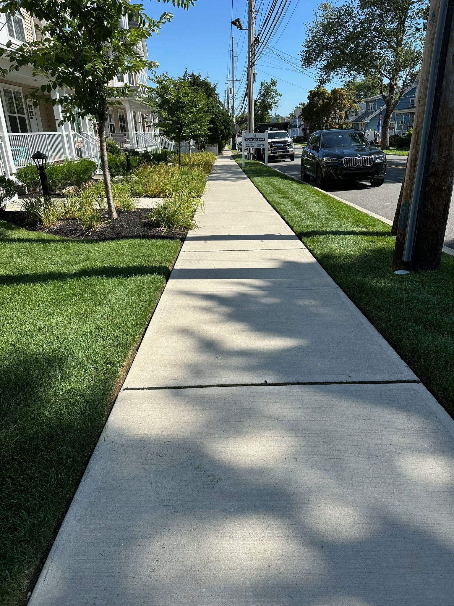 Sidewalk bordered by green grass and shrubs, trees and cars on a sunny day.