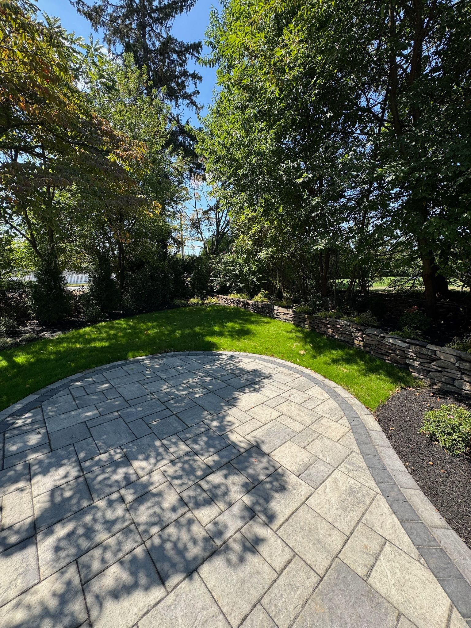 Stone patio bordered by grass and dark mulch, trees in background under blue sky.