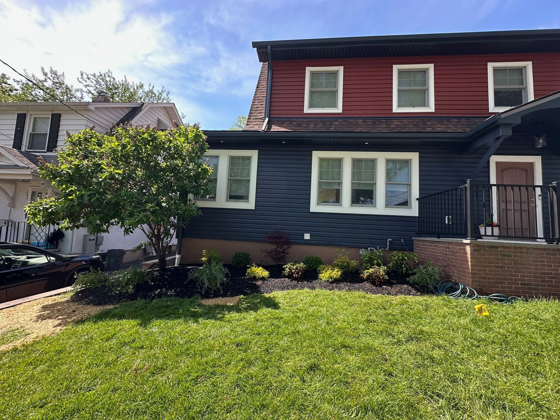 Two-story house with dark blue and red siding. Lush green lawn with flower beds and a tree in front.