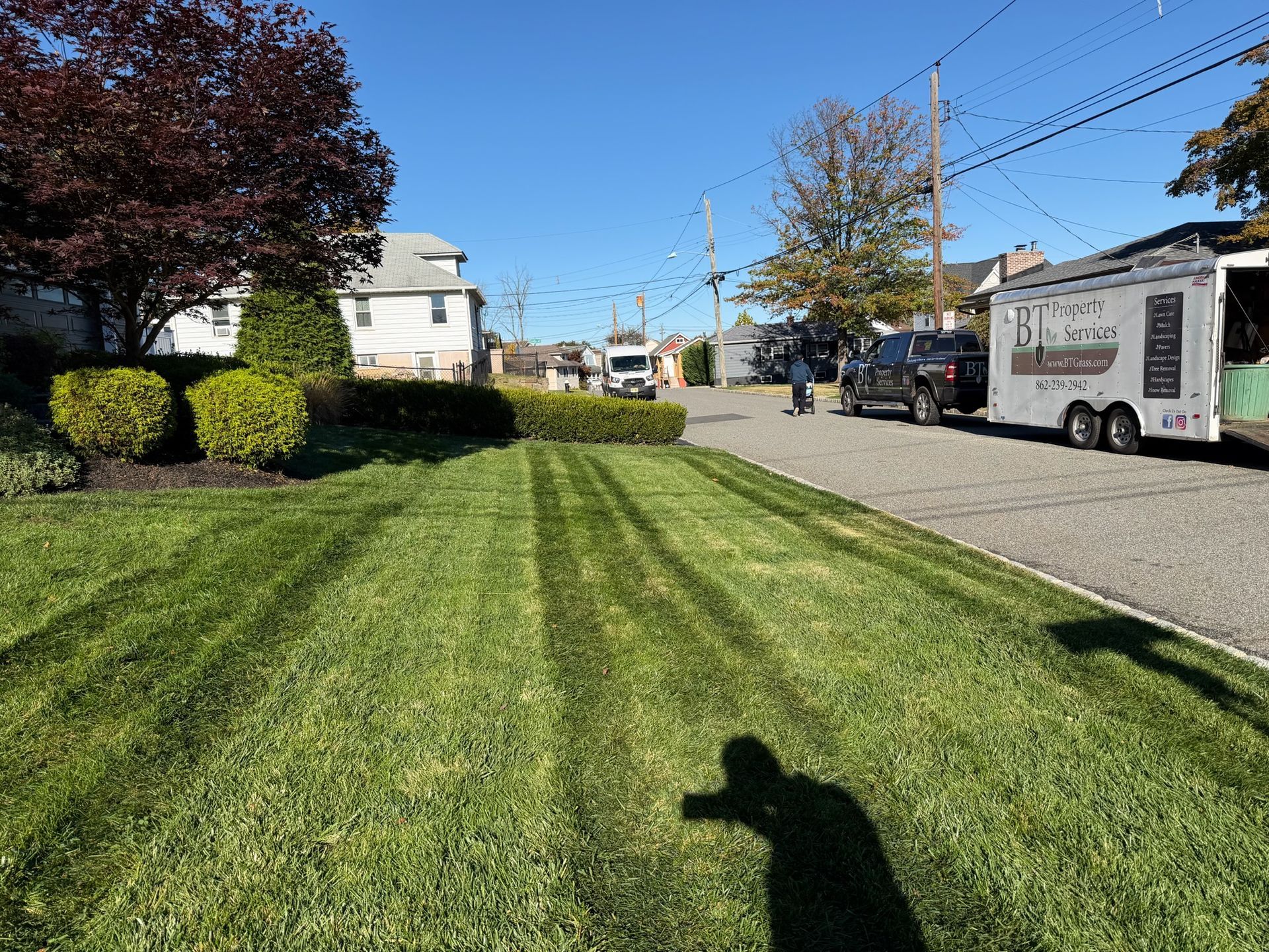 Lawn with freshly cut stripes; a person, truck, trailer, and houses are in the background on a sunny day.