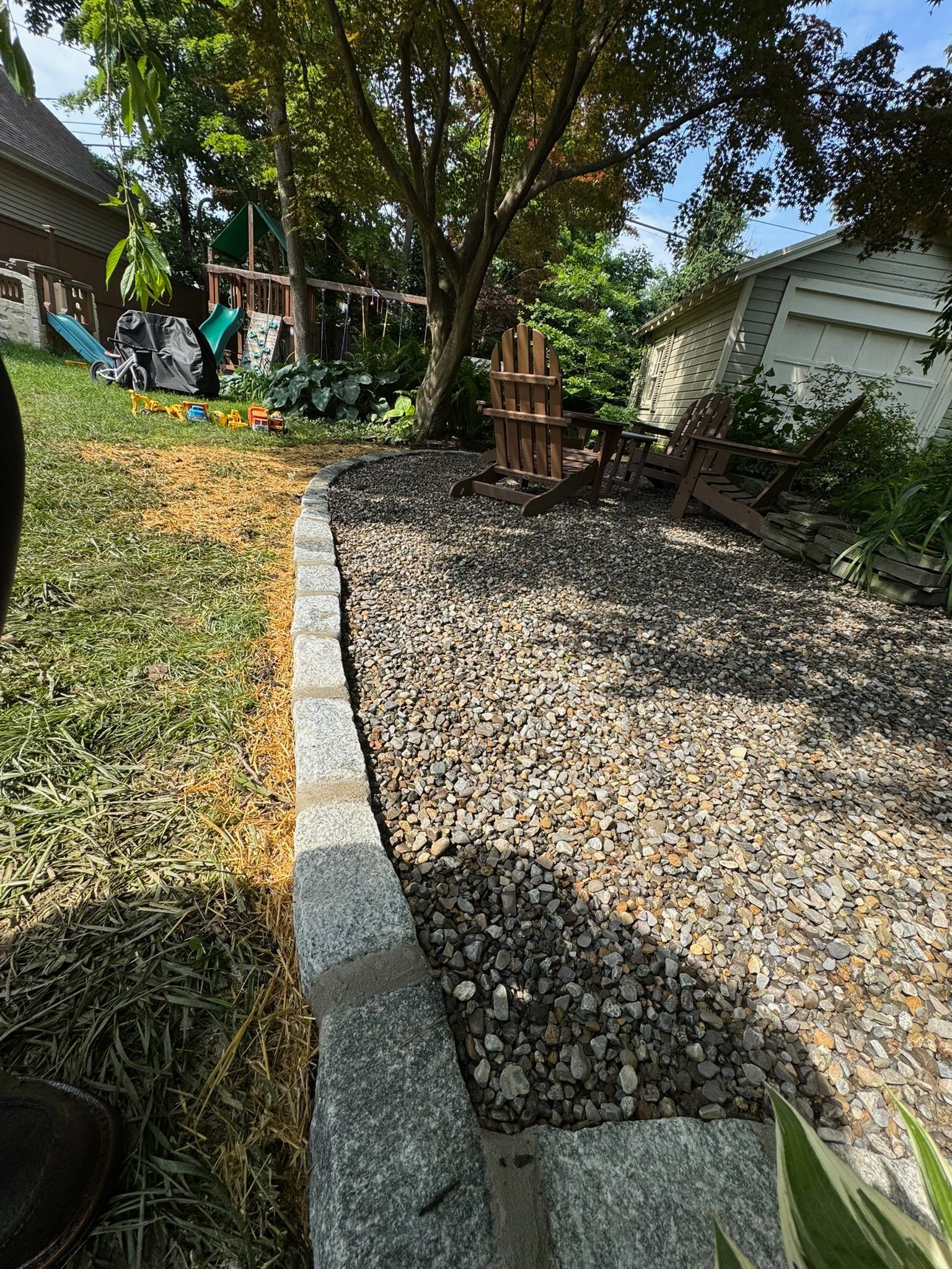 A backyard garden path of gravel, bordered by a low stone wall, with a tree in the center.