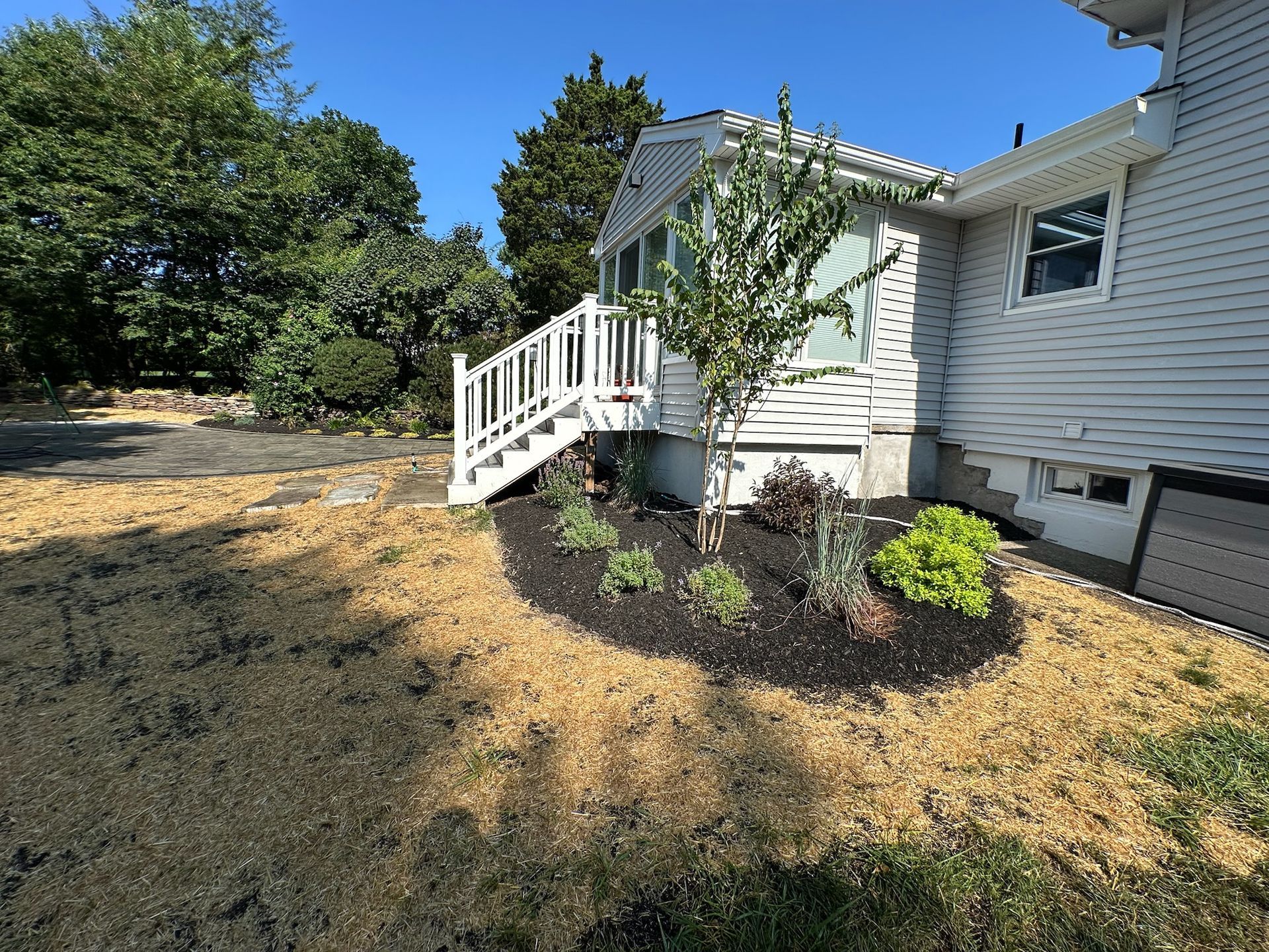 Side view of a house with white deck, new flower bed, and dry, brown yard.