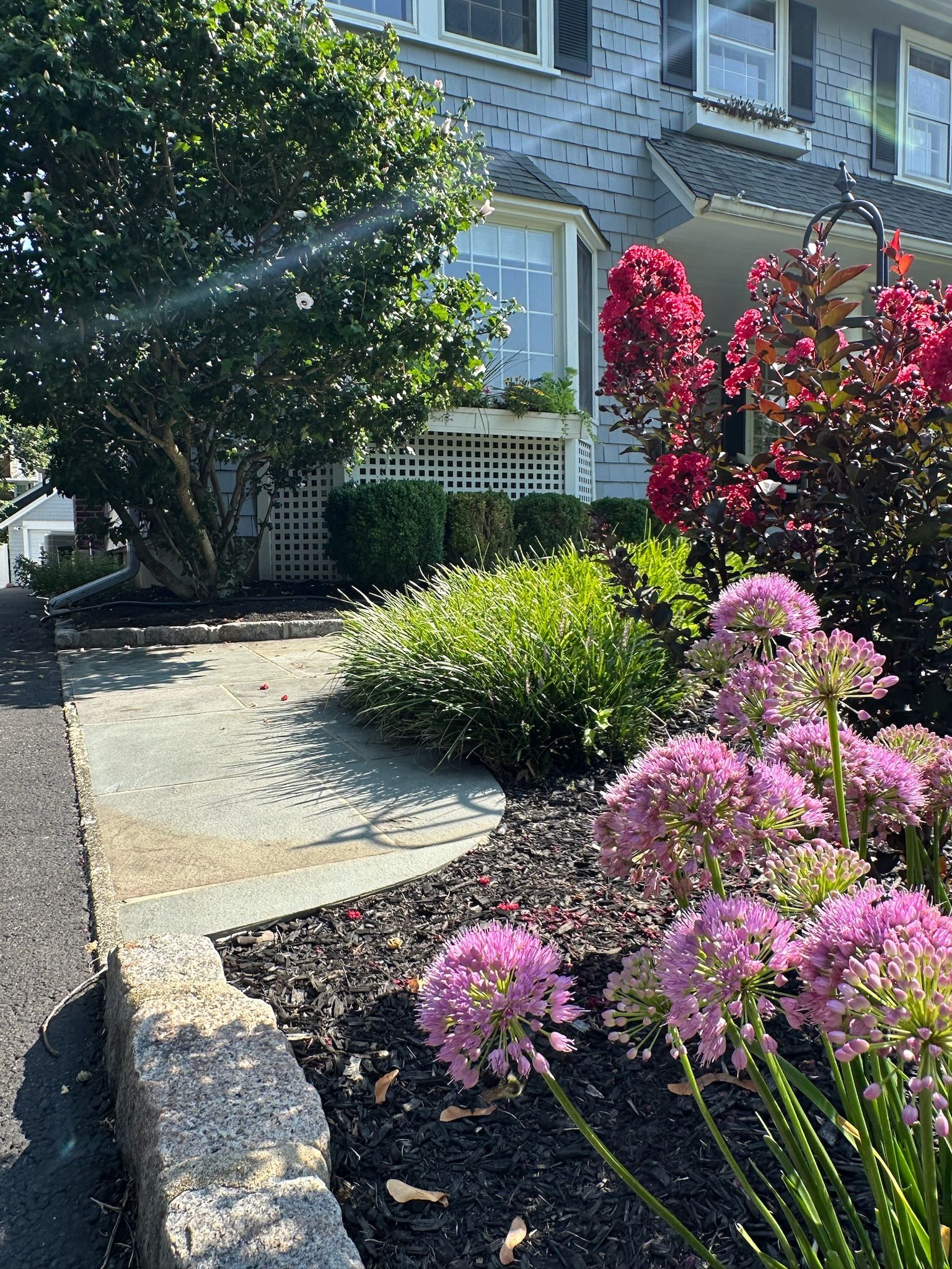 A stone path leads past a flower garden to a gray house with a bay window and dark shutters.
