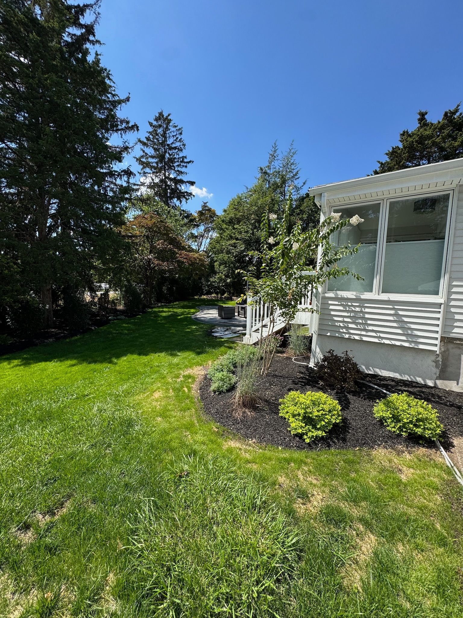 Green lawn with garden bed, white house, trees under blue sky.