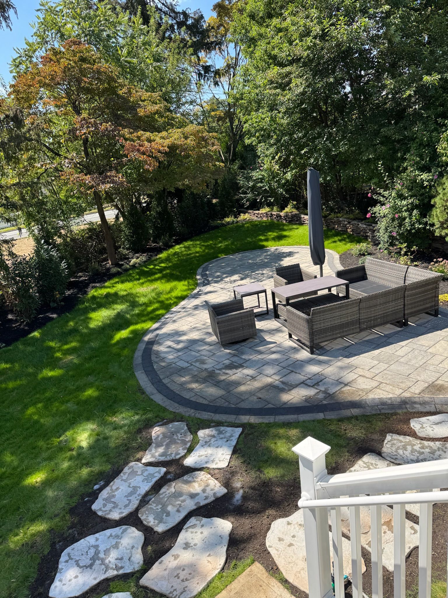 Patio with outdoor furniture, umbrella, and stone pathway surrounded by green grass and trees.