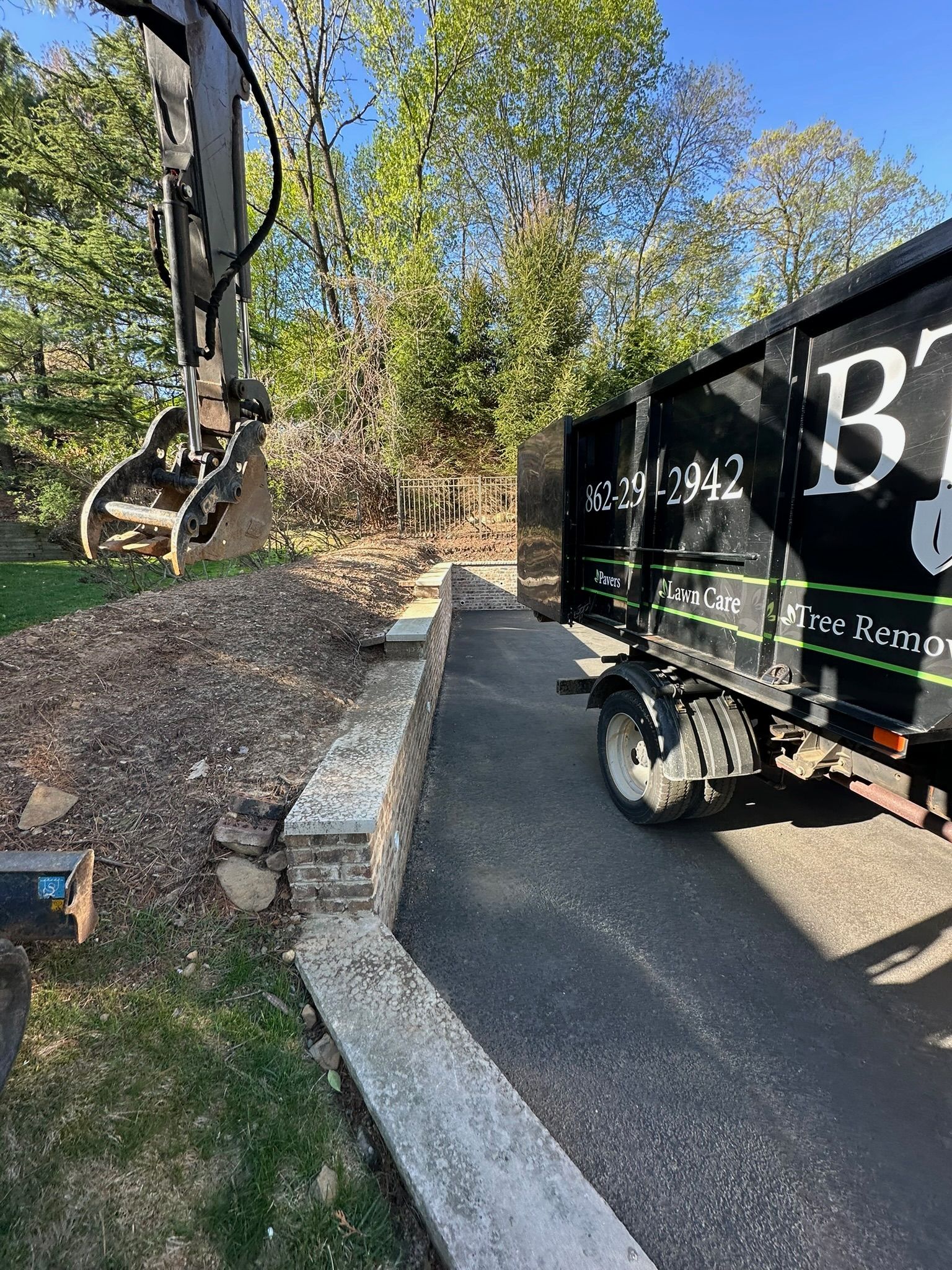 Excavator next to a black dumpster parked on a driveway, with a stone retaining wall in the foreground.