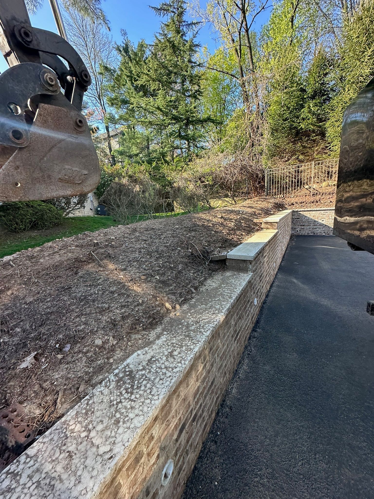 An excavator dumping mulch onto a pile next to a low concrete wall and paved driveway, in front of trees.
