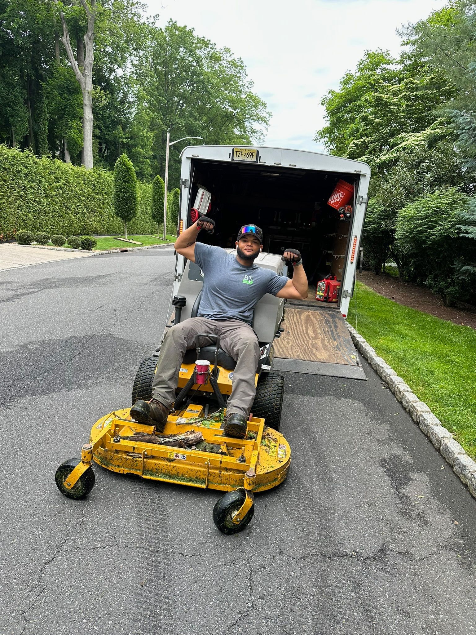 Person sitting on a yellow lawnmower, posing, with a truck in the background. Asphalt road and green foliage are visible.