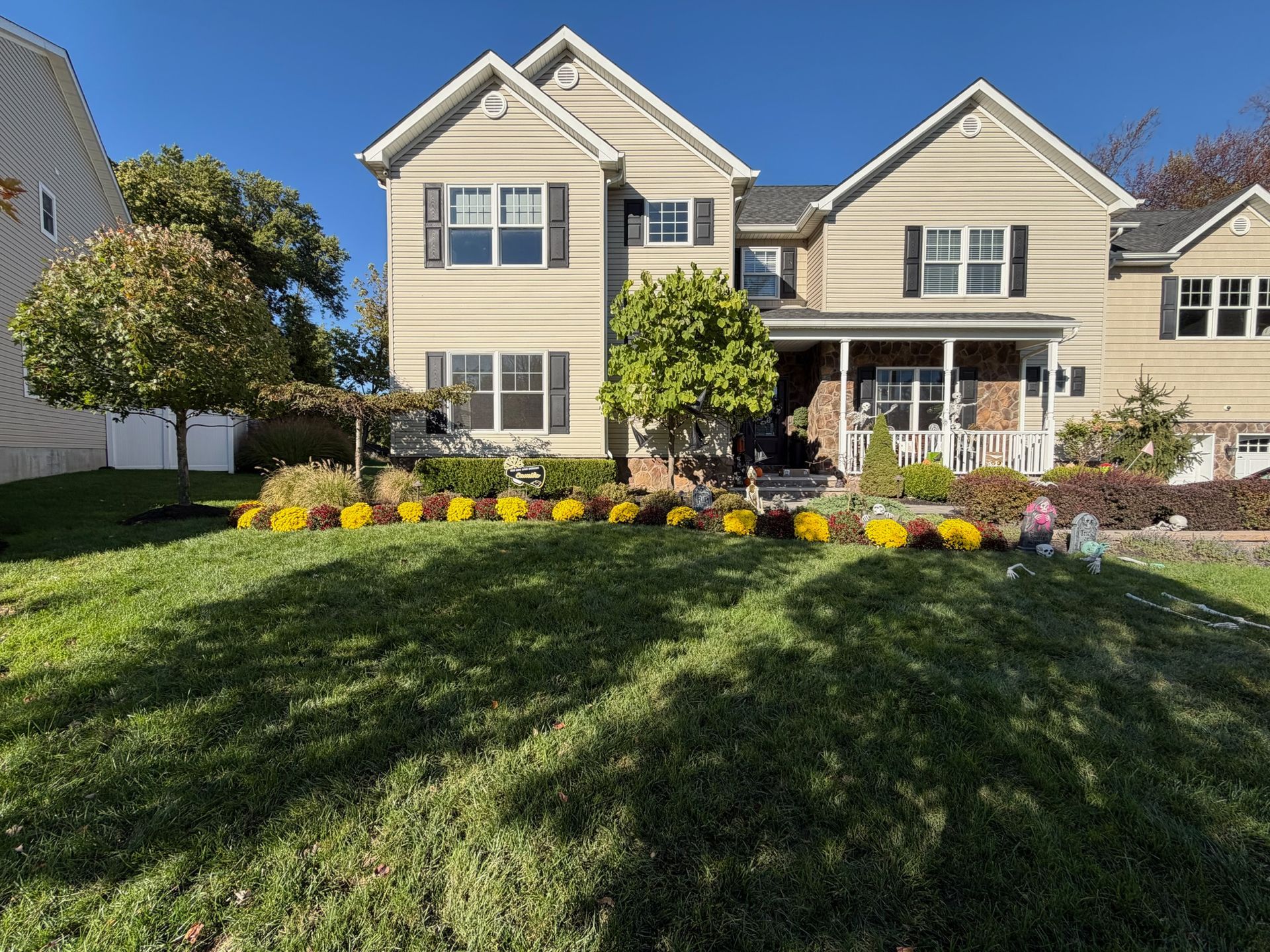 Two-story beige house with black shutters, yellow mums, and green lawn on a sunny day.