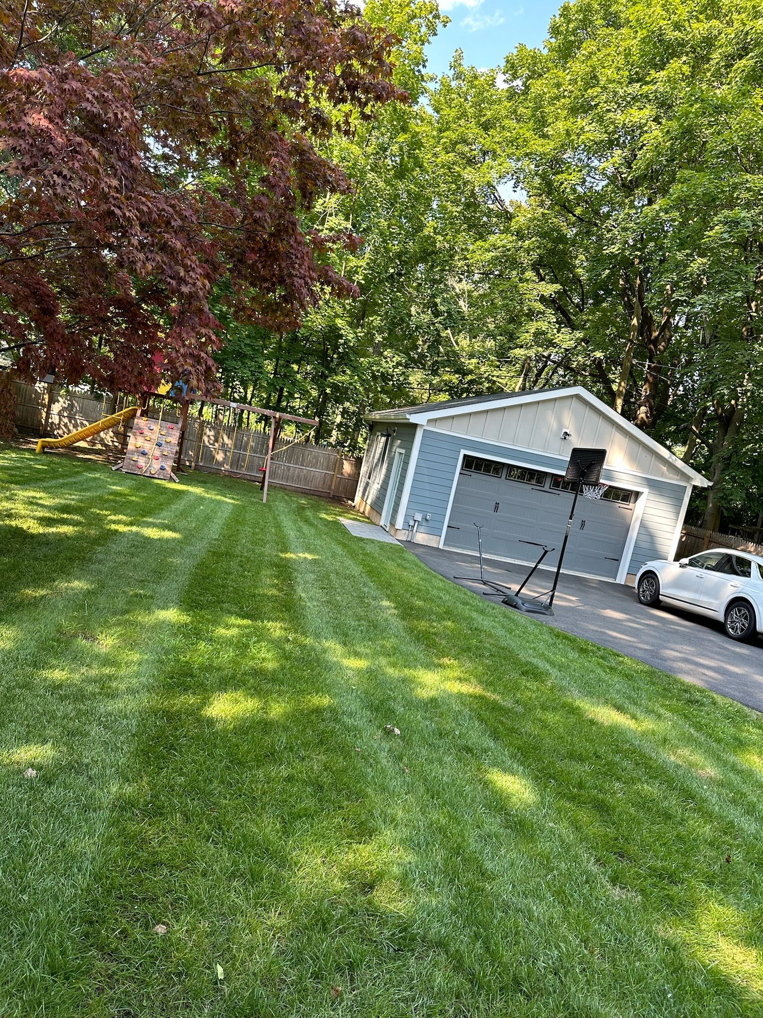 Green lawn with a two-car garage, a basketball hoop, and a playground set in the background.