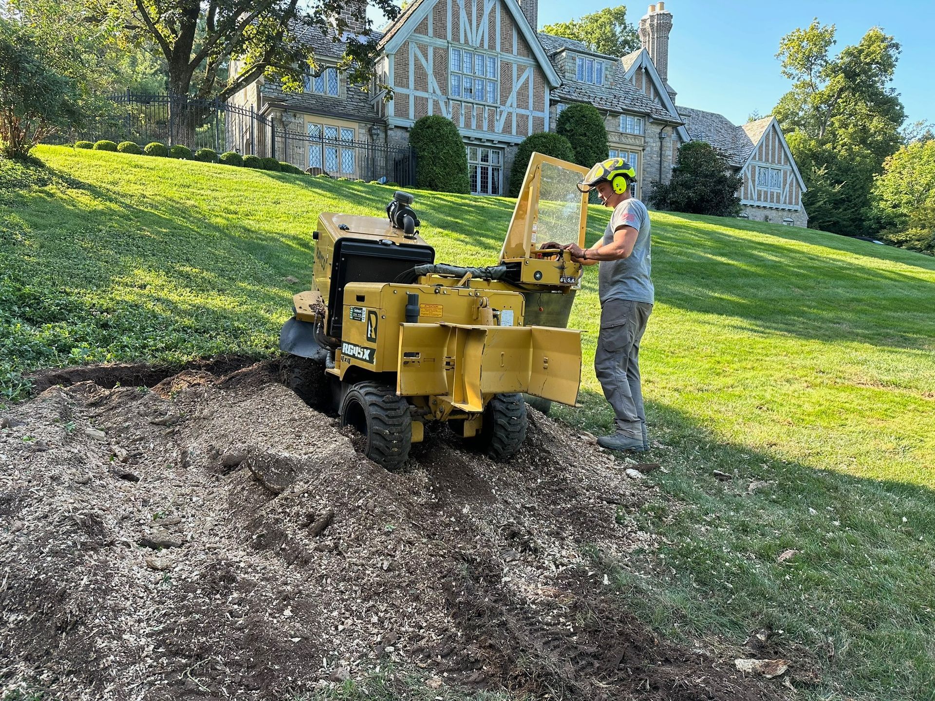 A person operating a yellow stump grinder on a grassy hill; large stone house in background.