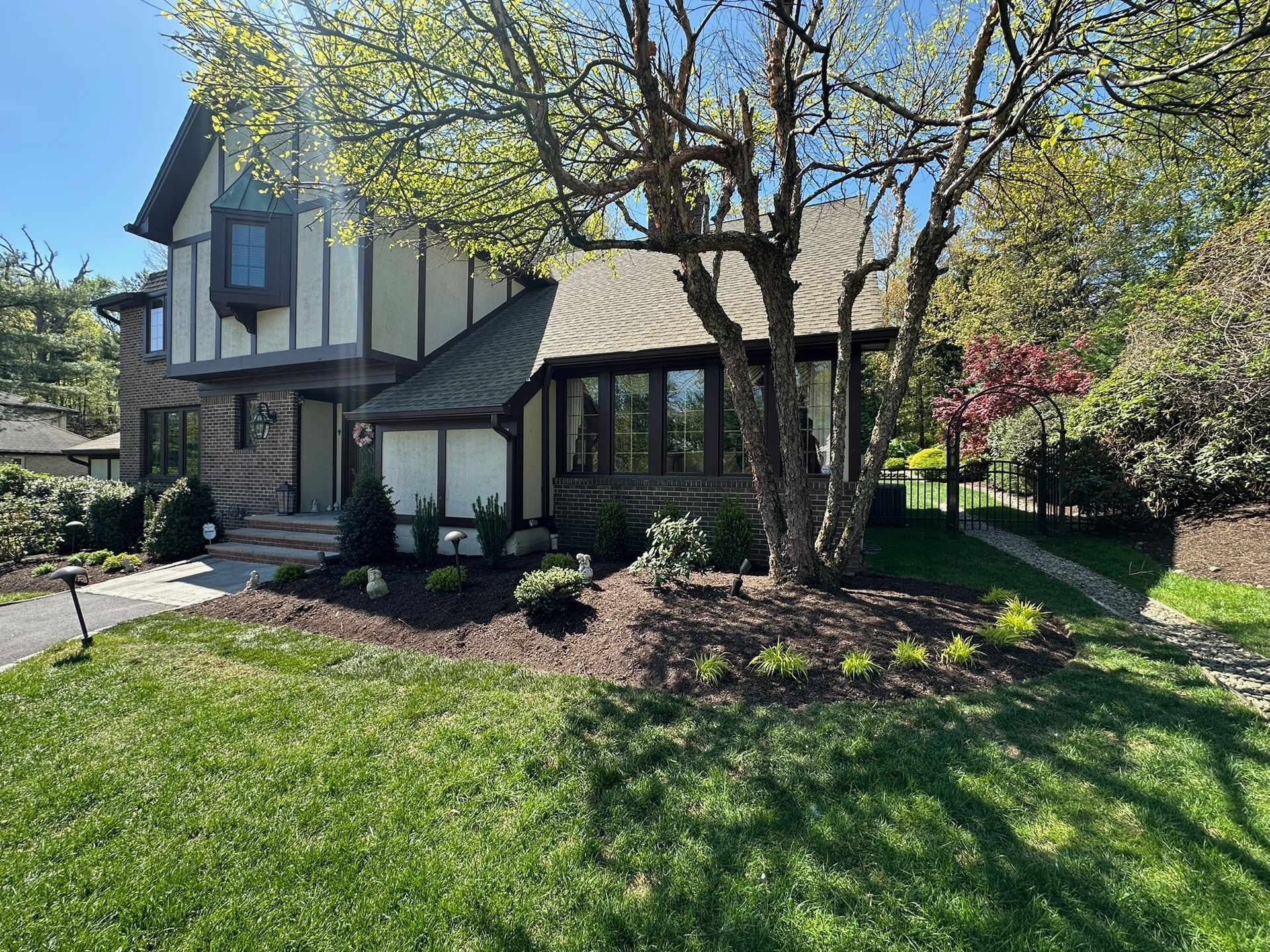 Tudor-style house with brown siding, a tree in front, and a green lawn on a sunny day.