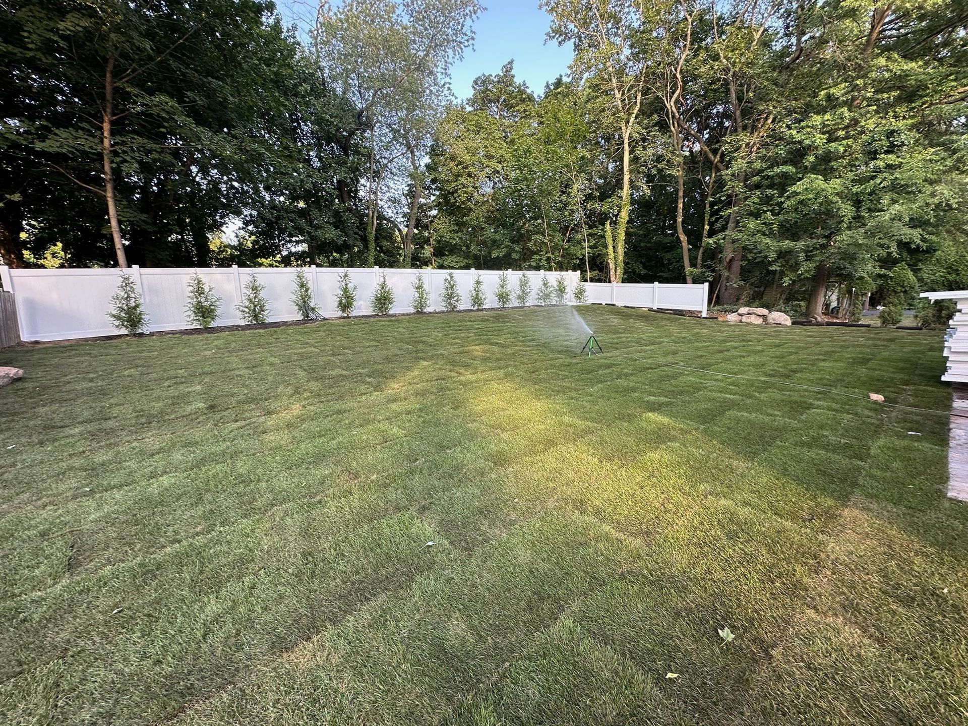 Green lawn with white fence, small trees, and a sprinkler in operation, with trees in the background.