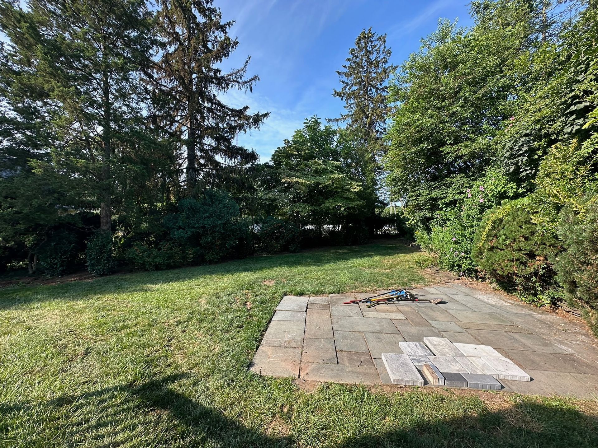 A grassy backyard with a stone patio, surrounded by trees under a blue sky.