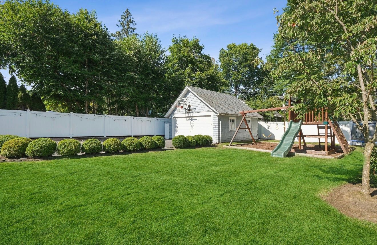 Green backyard with white fence, small shed, and wooden playset under blue sky.