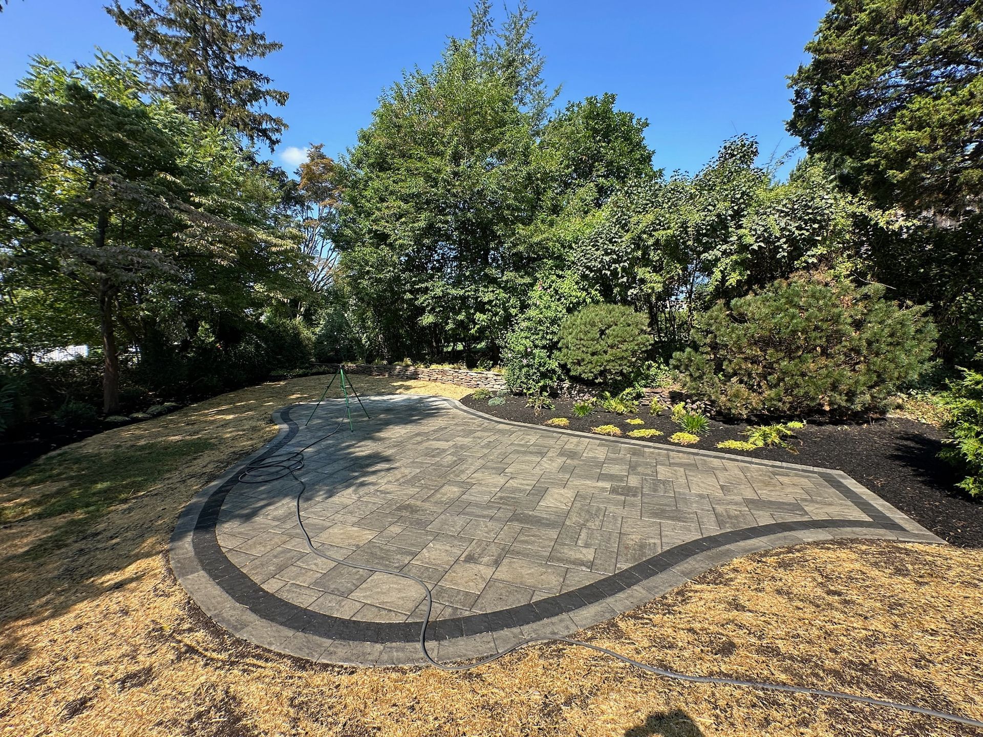 Paved patio bordered by dark edging surrounded by grass and various green shrubs under a blue sky.