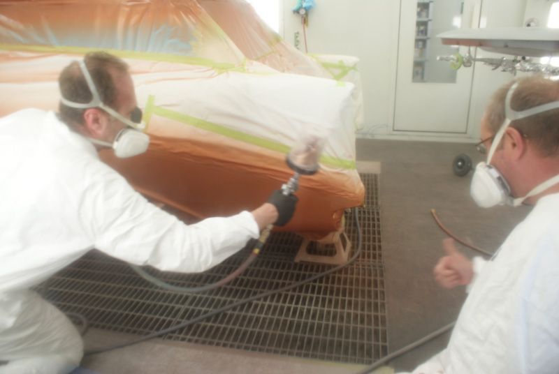 Two men wearing masks are painting a car in a garage.