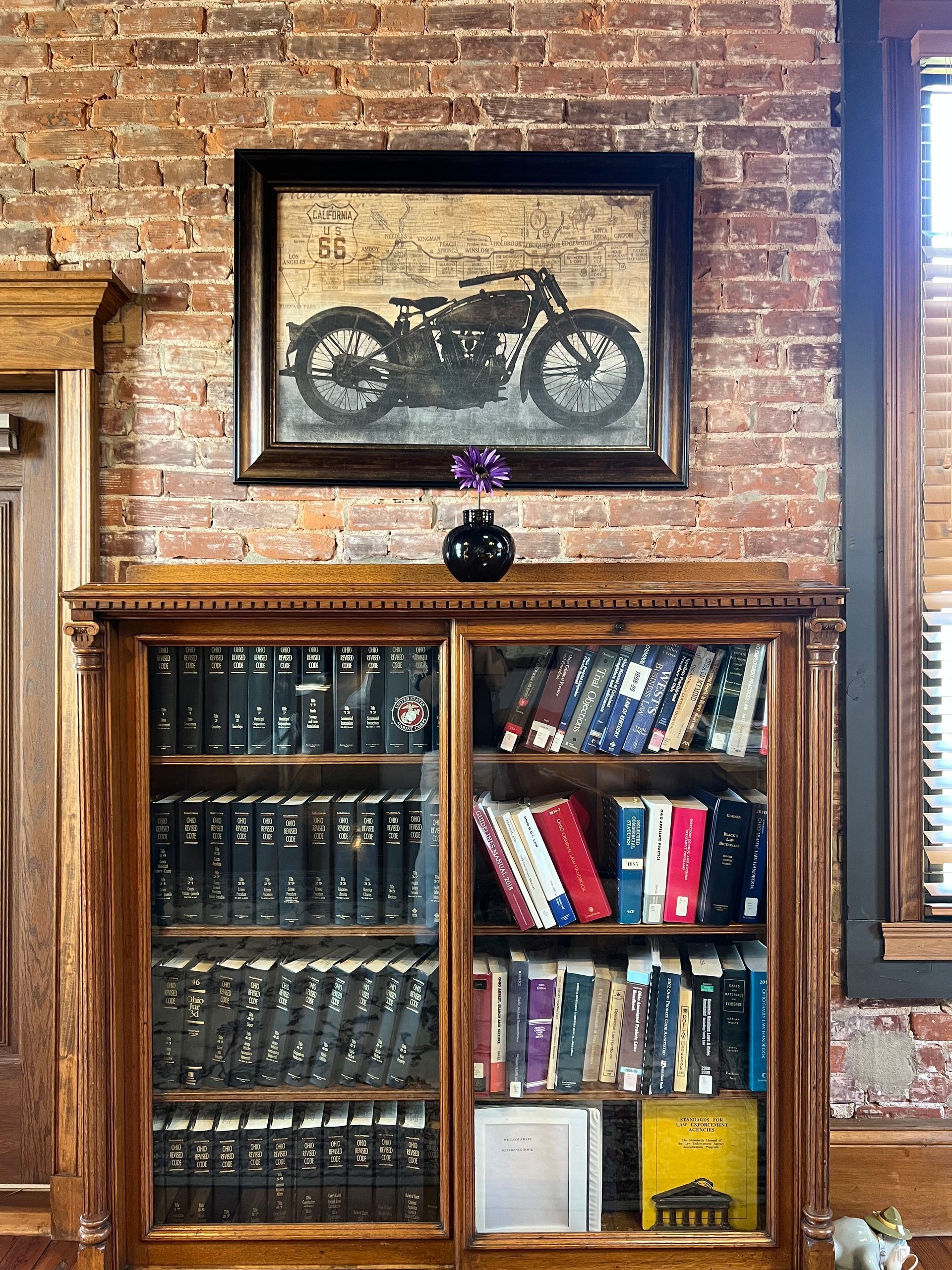 Brick wall with framed motorcycle art above a bookshelf filled with books and a small vase of flowers.