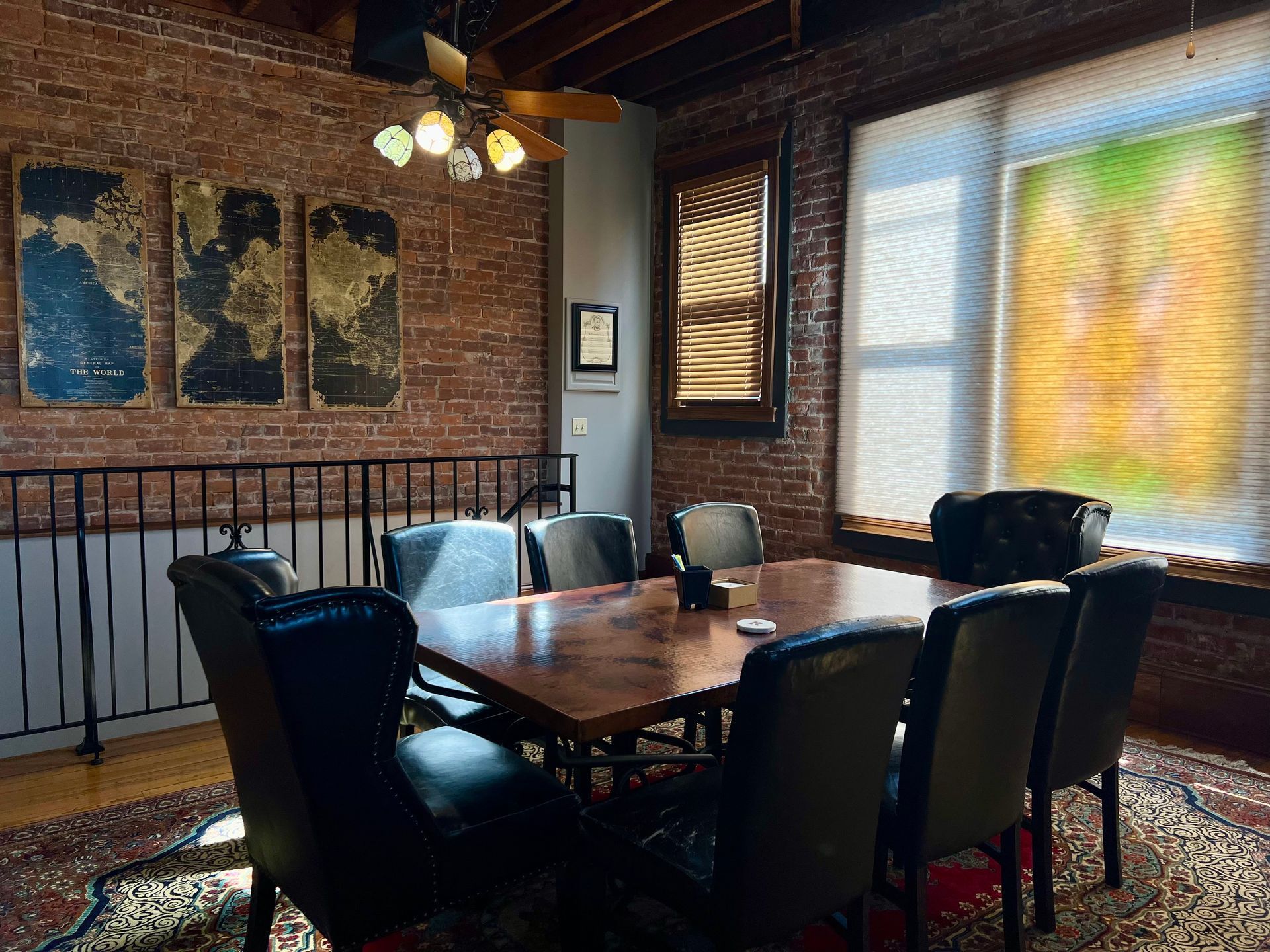 Dining room with a wooden table, black chairs, brick wall art, and bright window blinds.