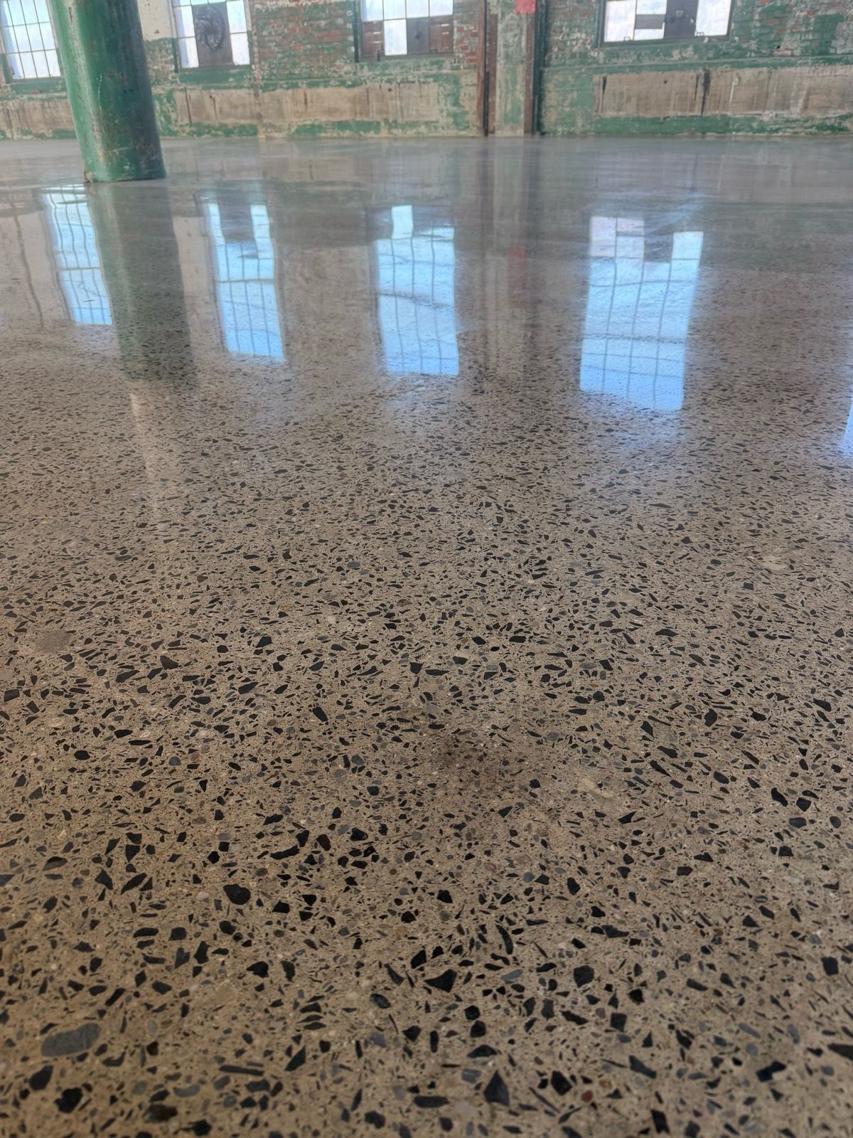 A high-gloss polished concrete floor with visible aggregate reflects industrial windows in a large, open room with green pillars and brick walls.